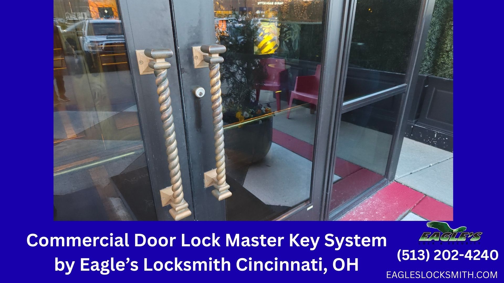 Close-up of a high-security commercial mortise lock on a metal front door in Cincinnati. The lock is etched with "EAGLE'S LOCKSMITH CINCINNATI." The blurred background shows the Cincinnati skyline and street activity. This lock is a candidate for conversion to a master key system.